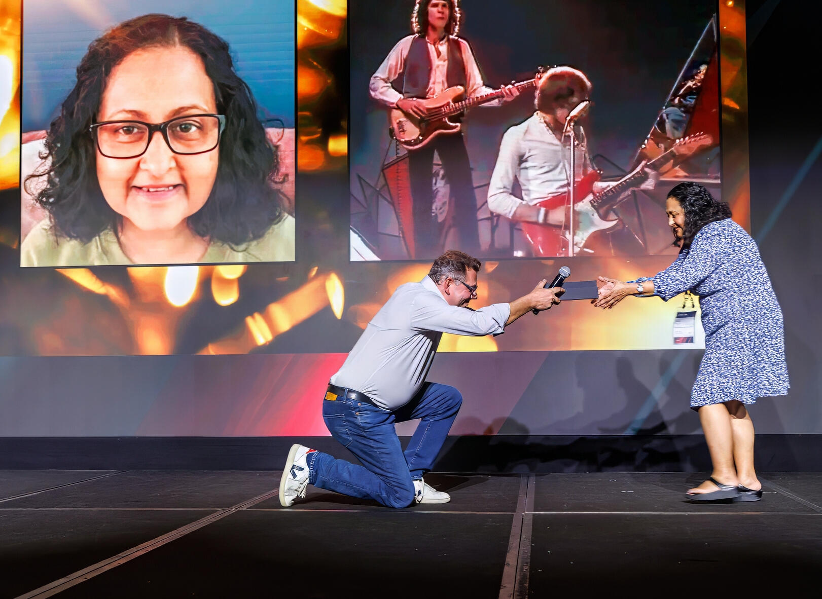 A memorable award handoff moment on stage during a corporate event in NYC, with a large screen backdrop displaying event visuals.